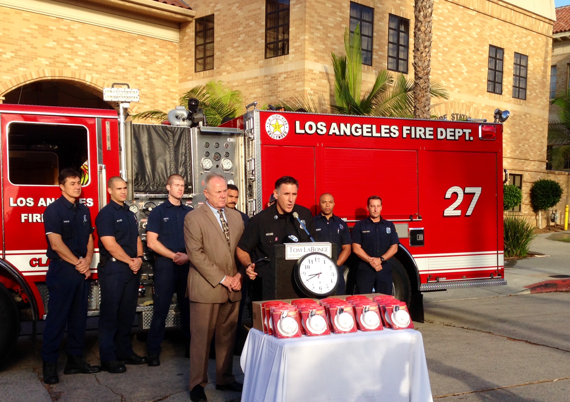 LAFD Captain Moore speaking at lectern. Firefighters and a fire engine in the background and a table of smoke alarms in the foreground.