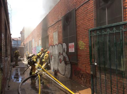 Looking down alley next to building, firefighters working to make entry and setting up hose lines