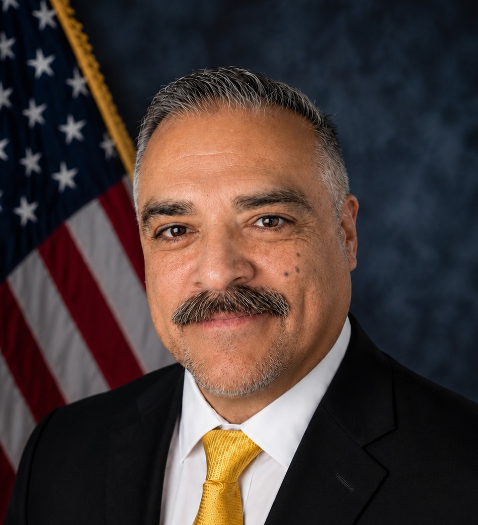 A formal head-and-shoulders portrait of a person wearing a black suit, white dress shirt, and bright yellow tie, smiling slightly at the camera. They have short gray hair, a neatly trimmed mustache and goatee, and are posed in front of a dark blue studio backdrop with a U.S. flag visible on the left side of the image. The lighting is even and professional, typical of an official or organizational portrait.