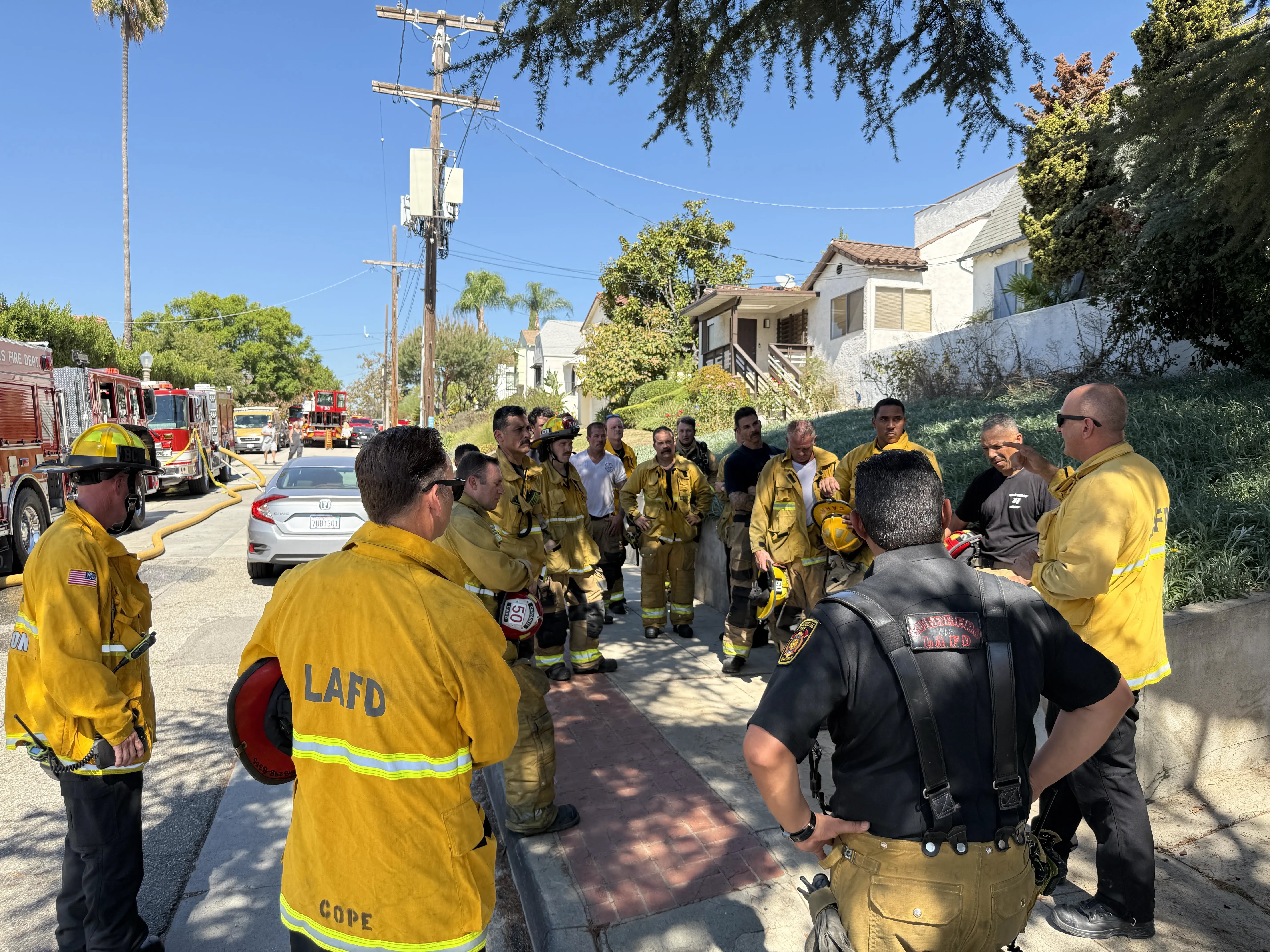 LAFD personnel in turnouts and uniforms gather outdoors on sidewalk for an operational briefing during incident.