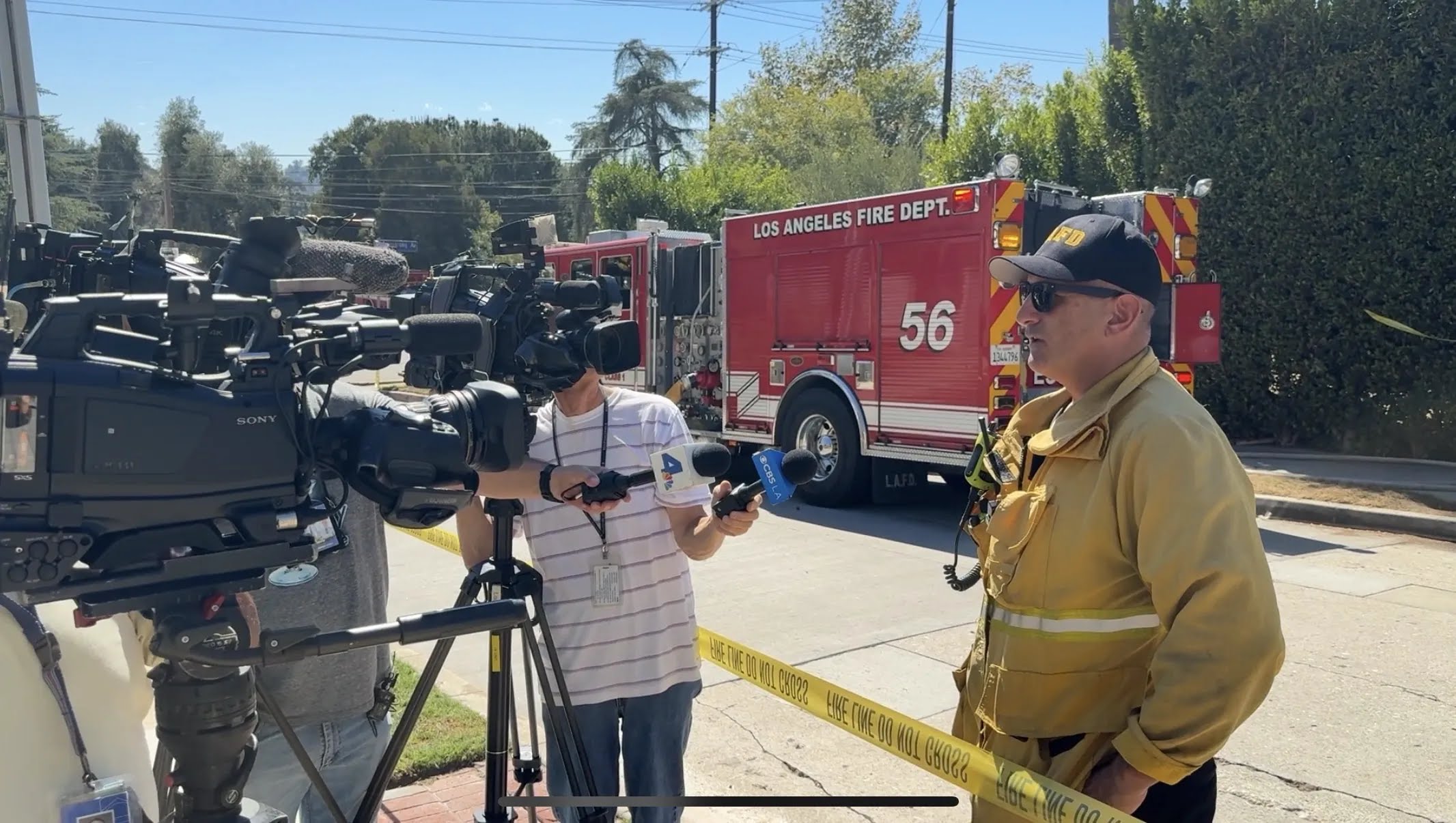 LAFD official speaks to news reporters holding microphones in front of Engine 56 during scene update.