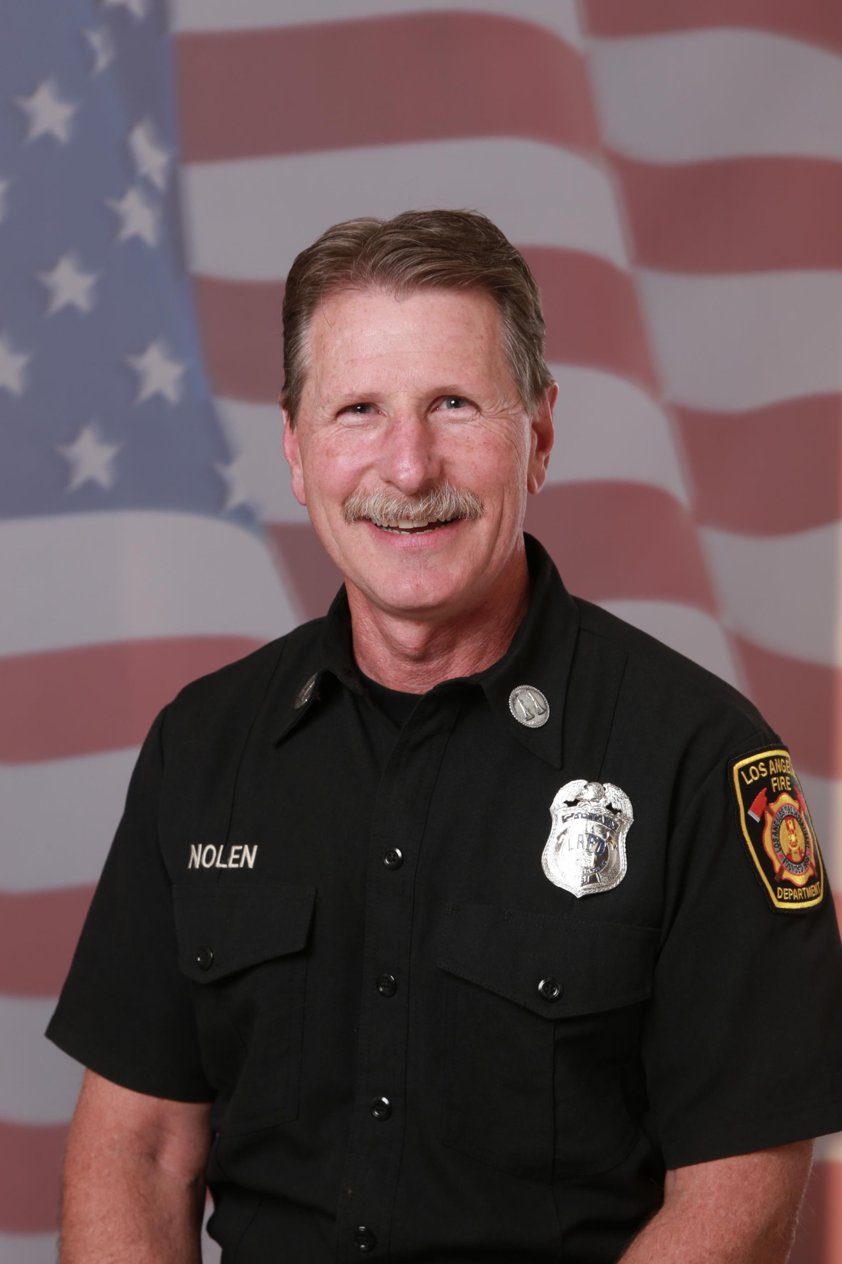 Portrait of a smiling man in a black firefighter uniform with patches, set against a blurred American flag background, conveying pride and professionalism.