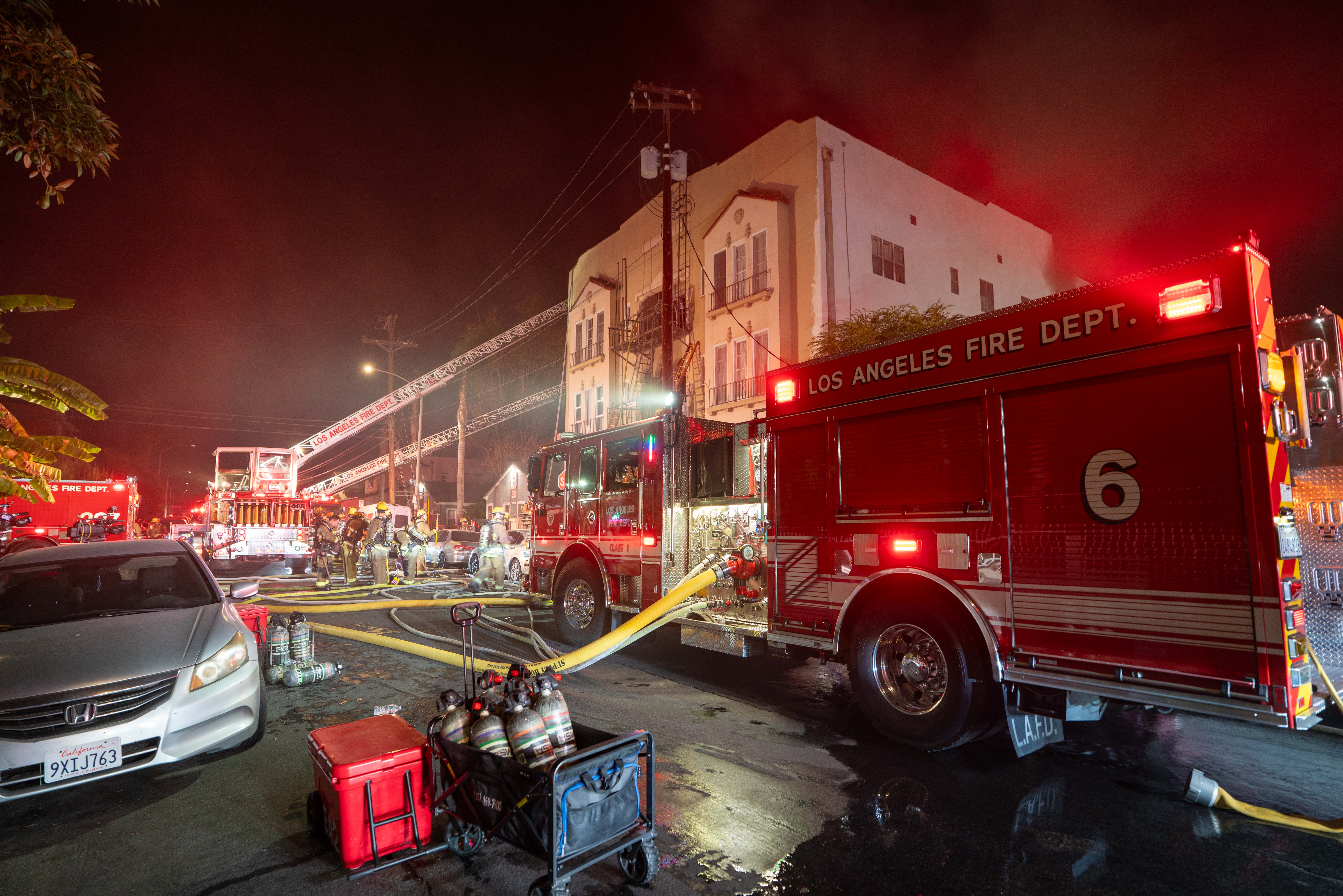 Los Angeles firefighters battle a nighttime blaze beside a red Fire Department engine, with hoses, equipment, flashing lights, and wet pavement.