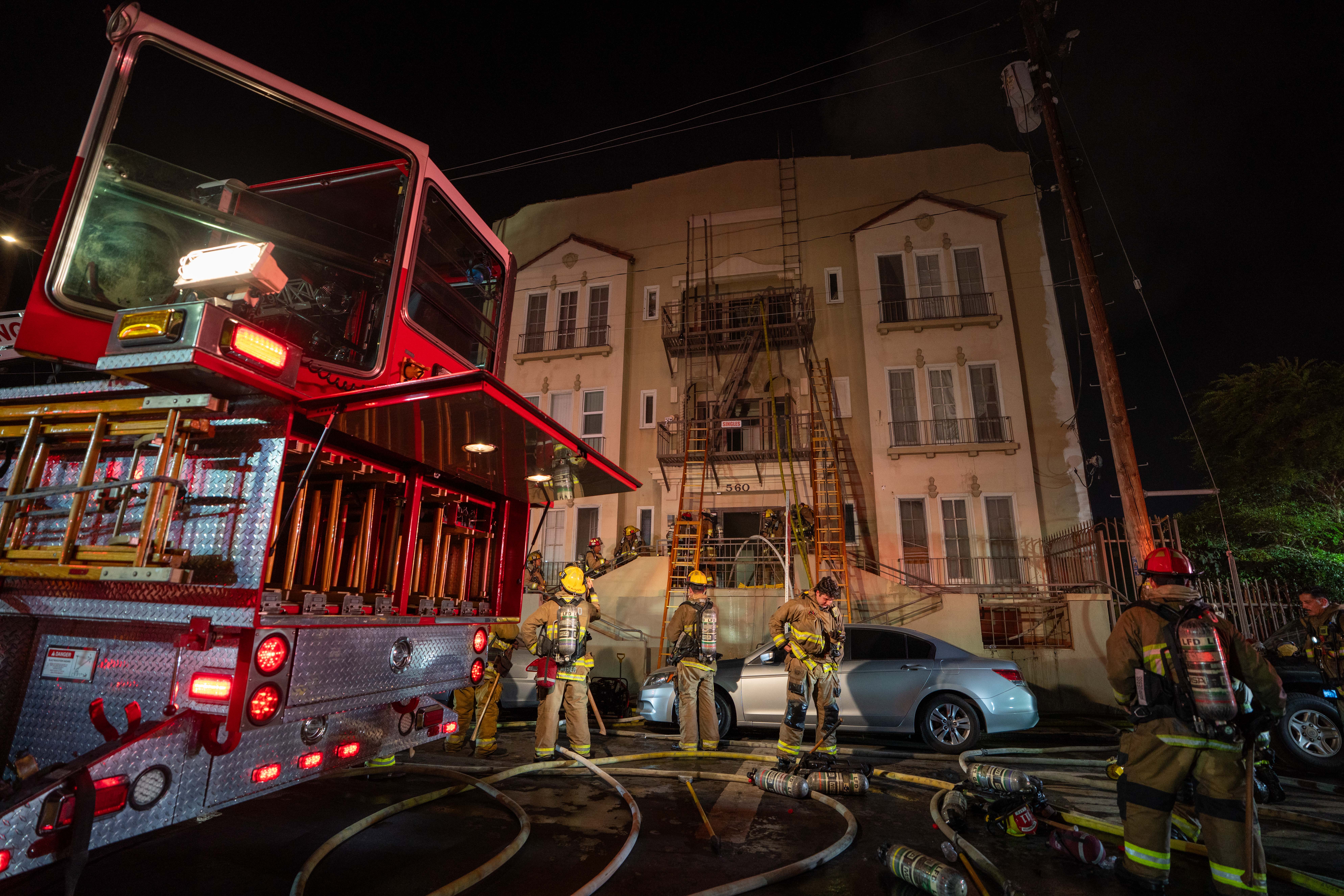 Firefighters work beside a red fire truck and hoses at night, responding outside a multi-story building; emergency lights cast a tense glow.