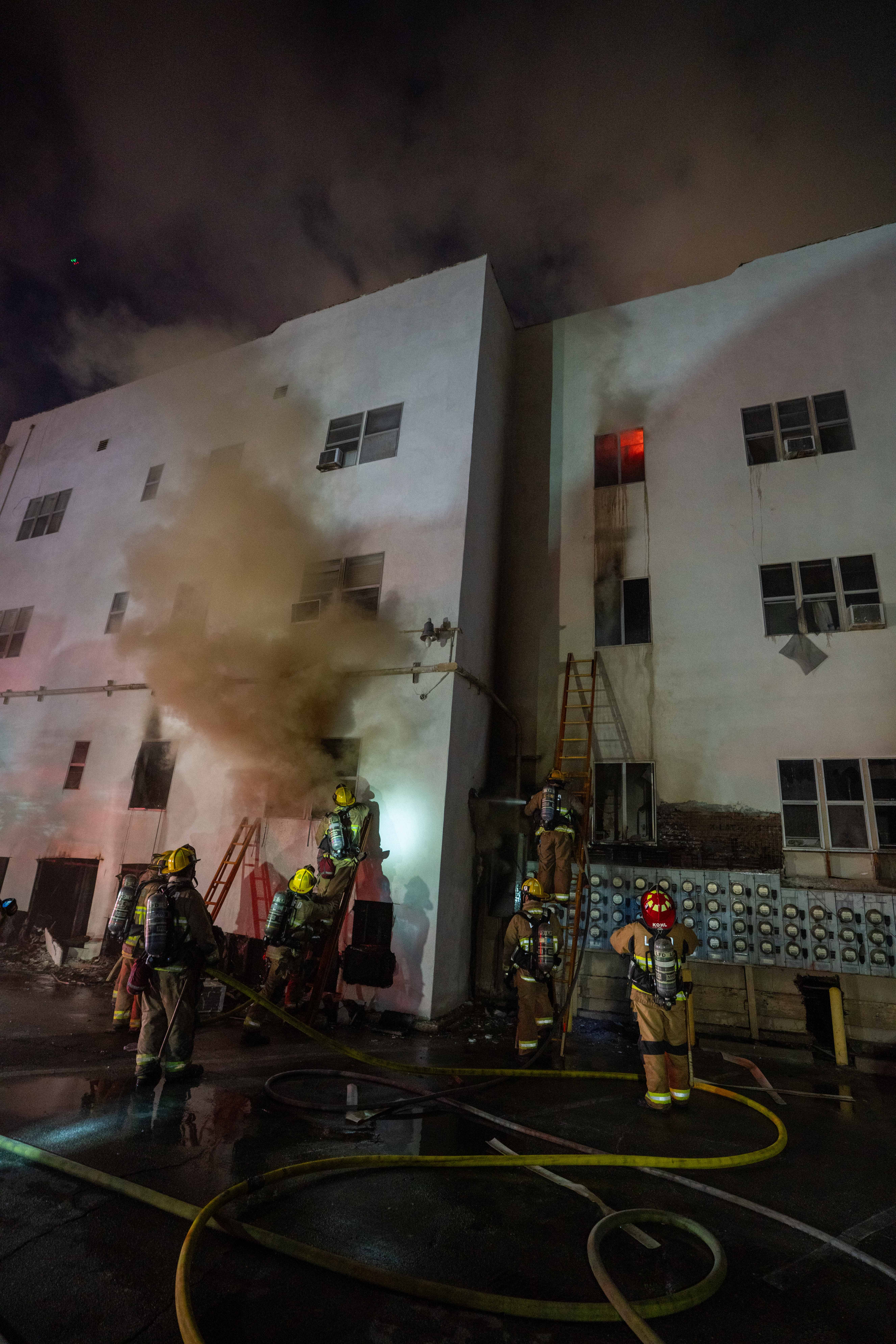 Firefighters in reflective gear battle a smoky nighttime blaze outside a multi-story building, with hoses across the wet ground and a window glowing red.