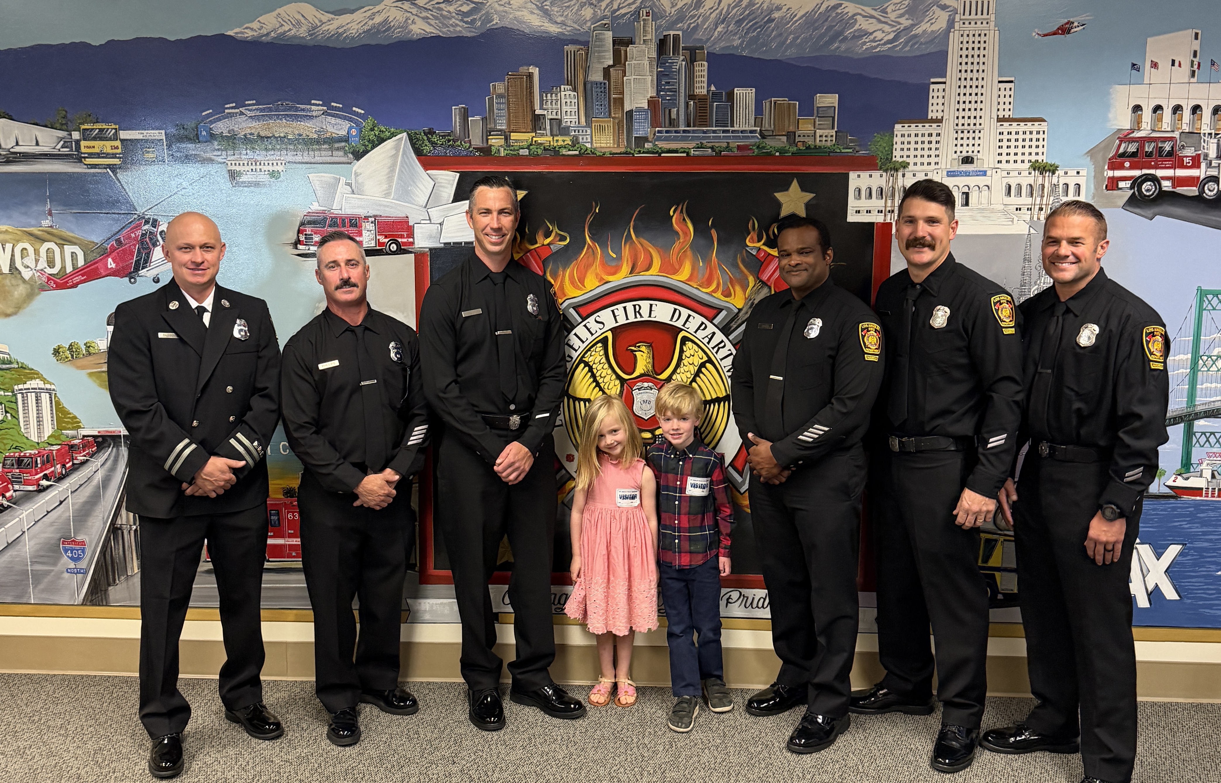 LAFD command staff and promoted personnel pose with children in front of a department mural at a ceremony.