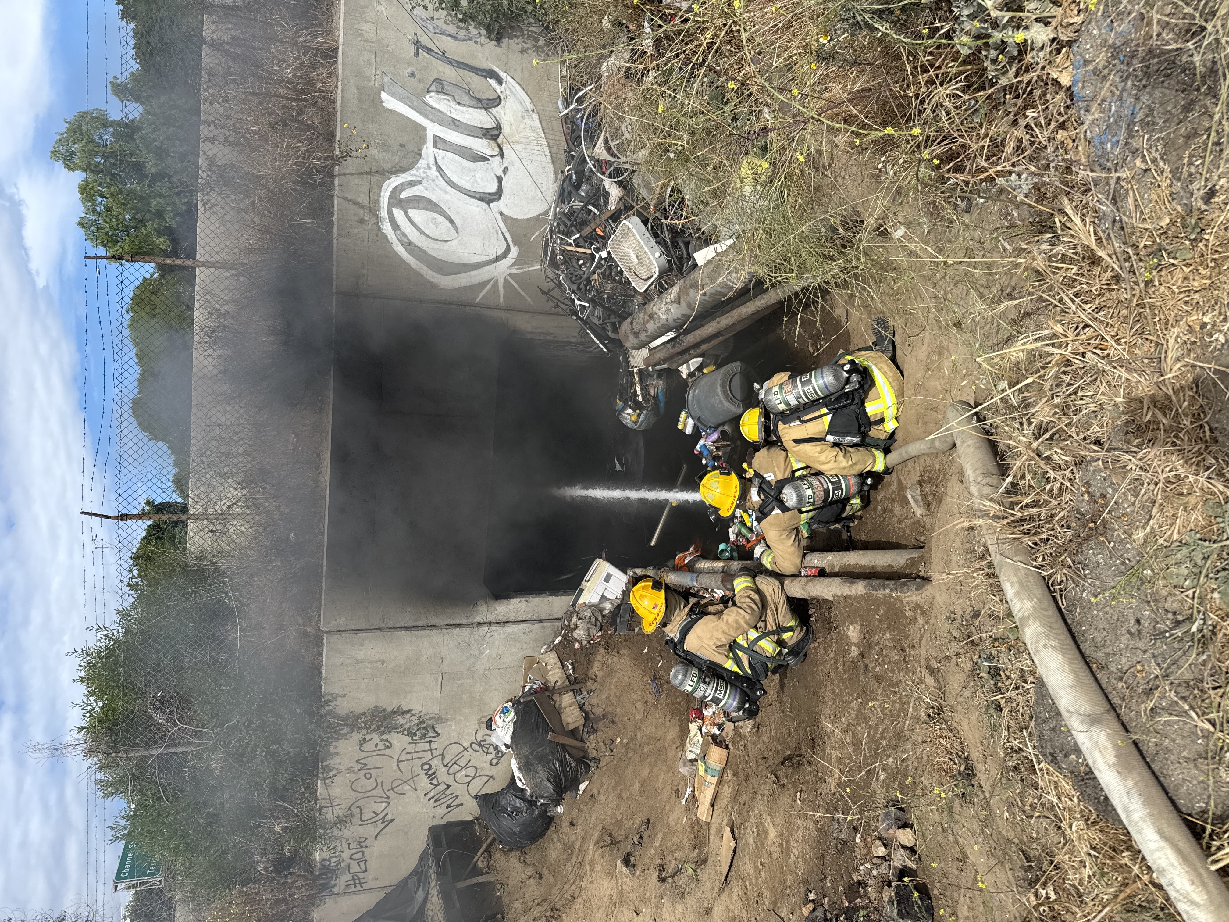 Firefighters with yellow helmets and gear spray water into a smoking access portal littered with debris. The scene is outdoors and suggests urgency.