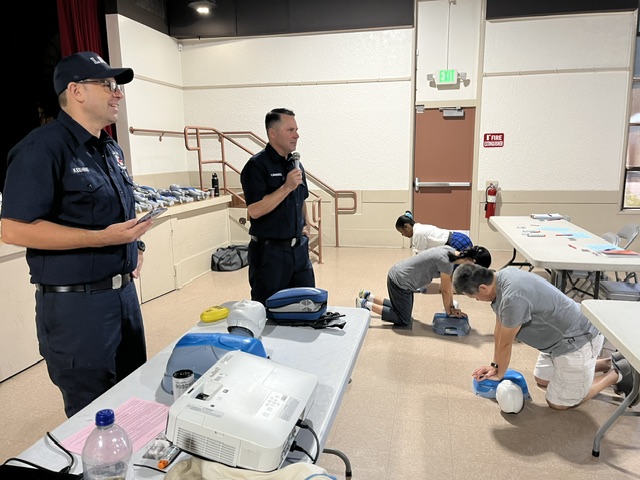 An LAFD instructor speaks into a microphone while another stands nearby, guiding community members kneeling on the floor to practice chest compressions on CPR manikins during a training session.