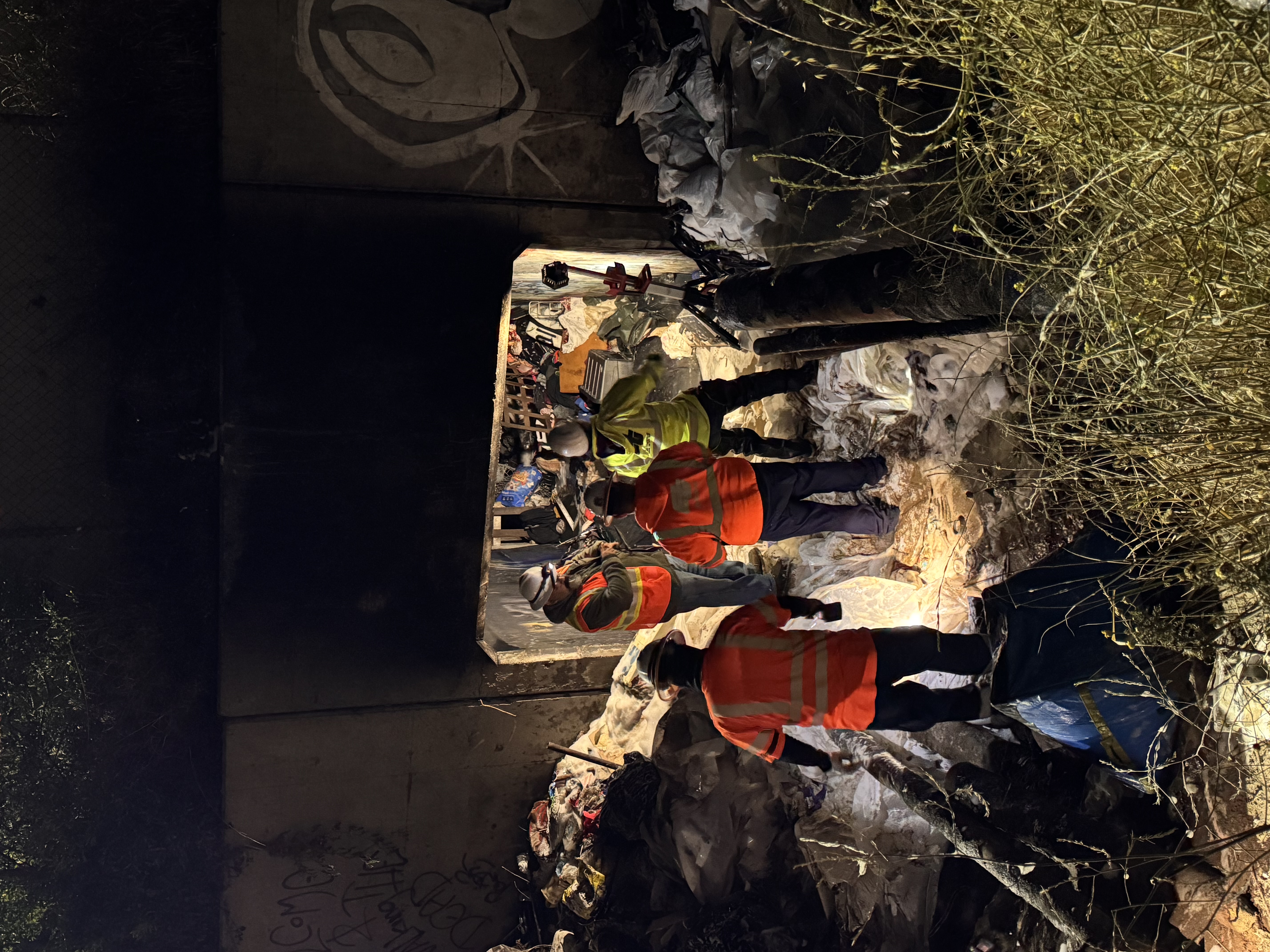 Four construction workers in reflective gear and helmets stand near a tunnel entrance filled with debris. The scene is dimly lit, creating a tense atmosphere.