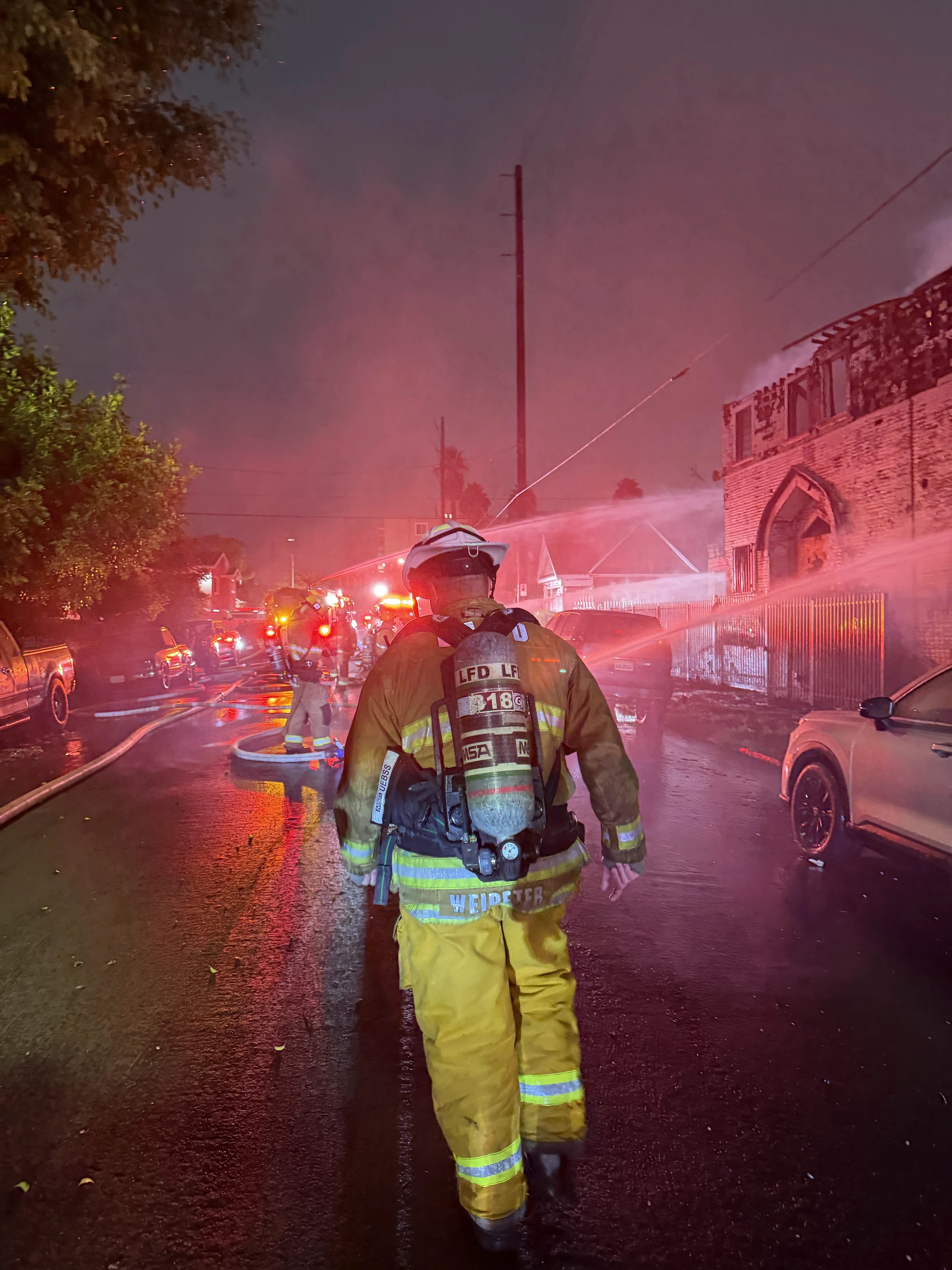 LAFD Chief Weireter walks toward flashing lights and hose streams at a major emergency structure fire wearing an SCBA air tank backpack with the number 18 on it