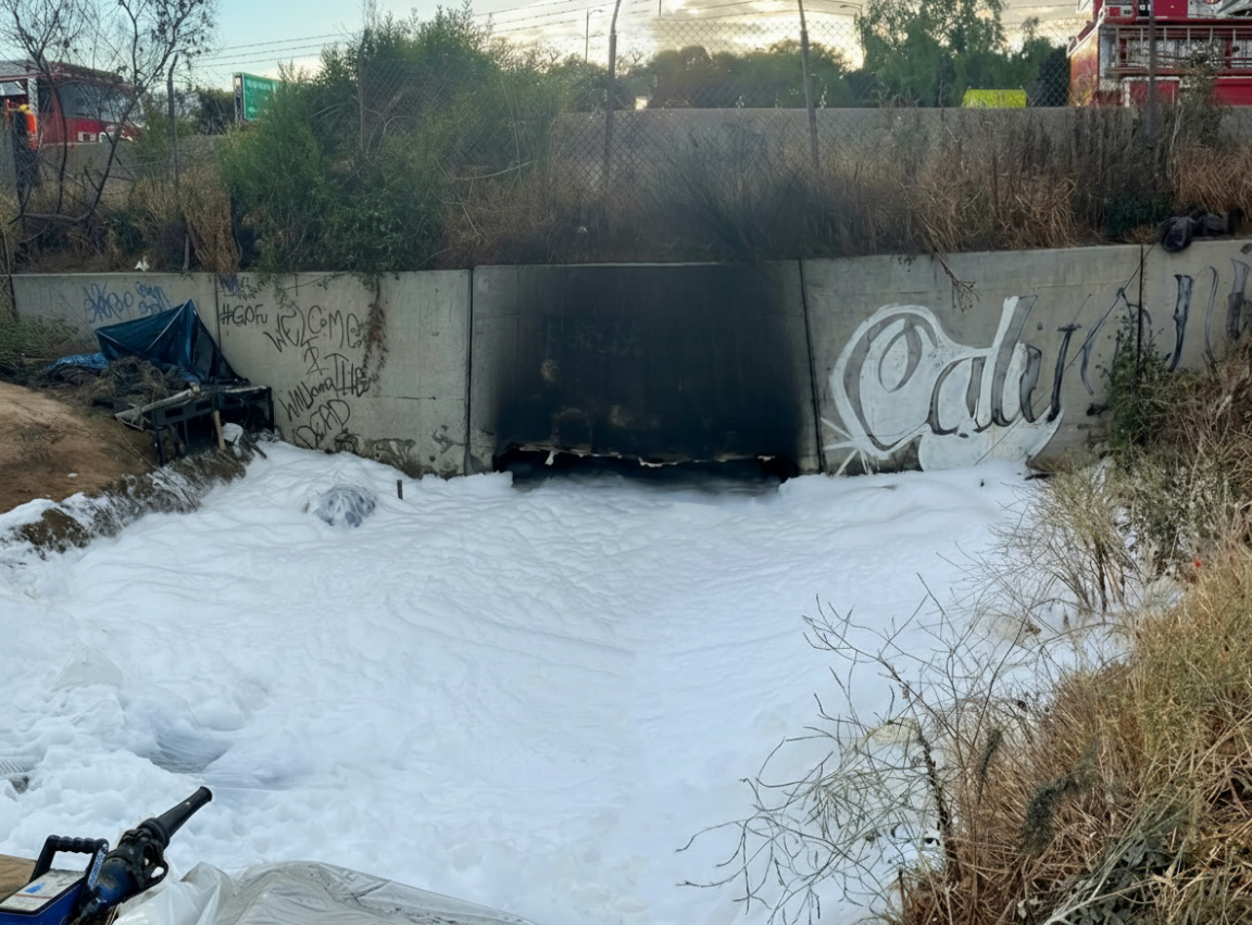 An opening in a highway embankment is filled with thick white foam. Surrounding concrete walls feature graffiti. Sparse vegetation and a fire truck are visible nearby.