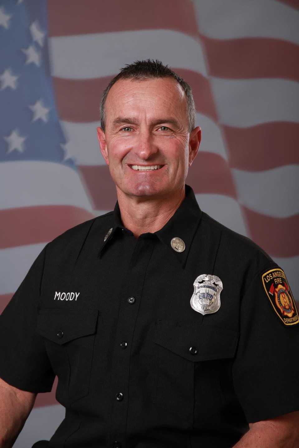 Formal Los Angeles Fire Department Portrait Photograph of LAFD Captain Richard Moody, smiling in his work uniform with badge, with a U.S. Flag motif in the background