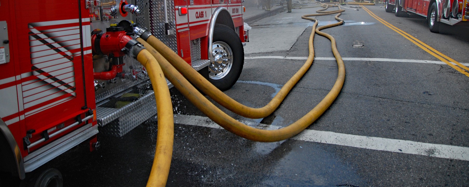 Yellow fire hoses snake from red fire engines, across a wet city street, conveying an urgent, focused emergency response.