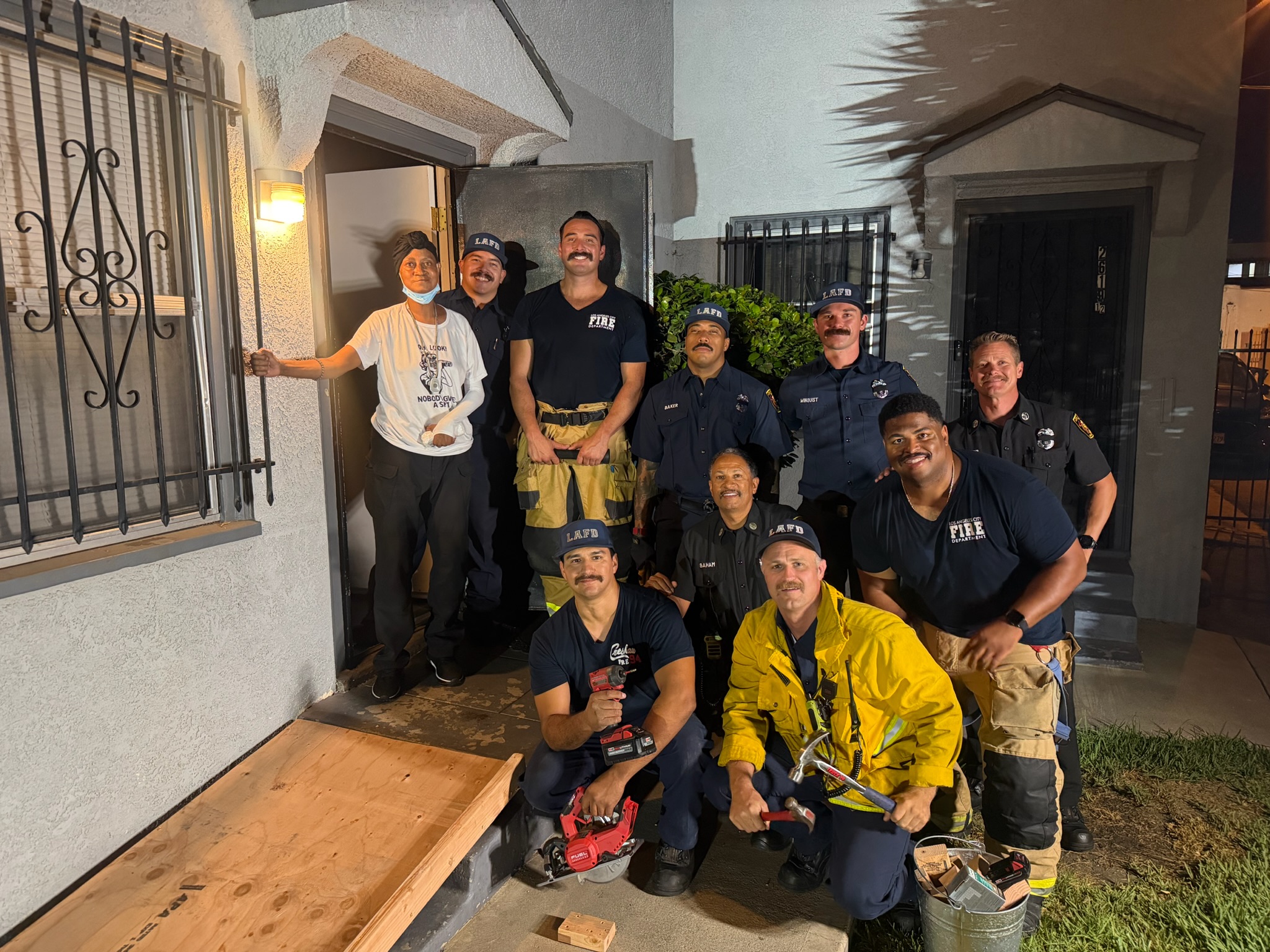 A group of Los Angeles Fire Department firefighters pose for a nighttime photo outside a home alongside the homeowner, following the installation of a custom wooden wheelchair ramp over the entrance steps.