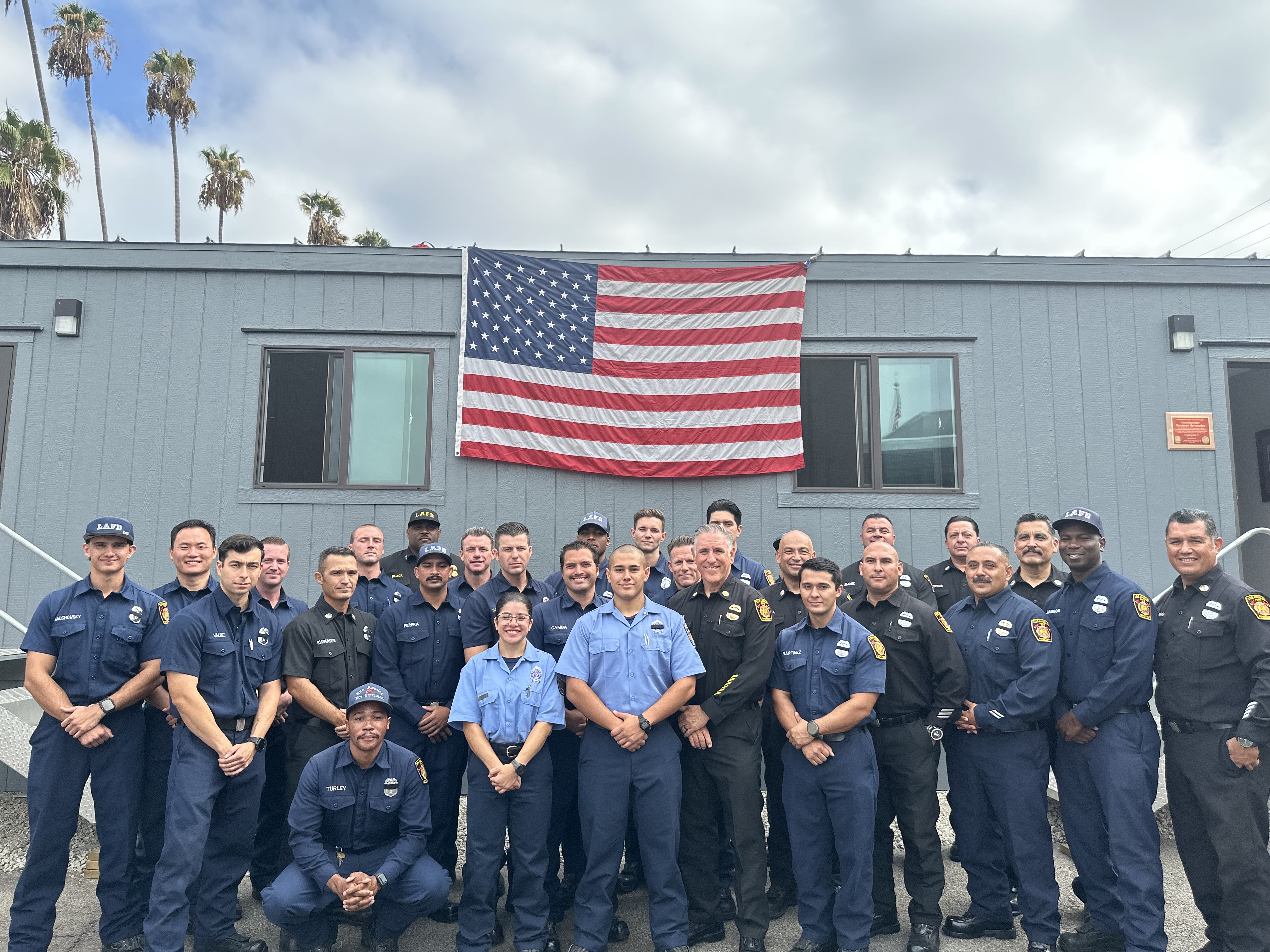 A large group of Los Angeles Fire Department (LAFD) personnel poses together for a group photo outdoors in front of a gray, single-story modular building.The group consists of over twenty individuals dressed in official dark blue and light blue LAFD uniforms. They are arranged in two rows, with one individual kneeling in the front left. A large American flag hangs prominently on the building's exterior wall directly behind the center of the group, positioned between two windows. 