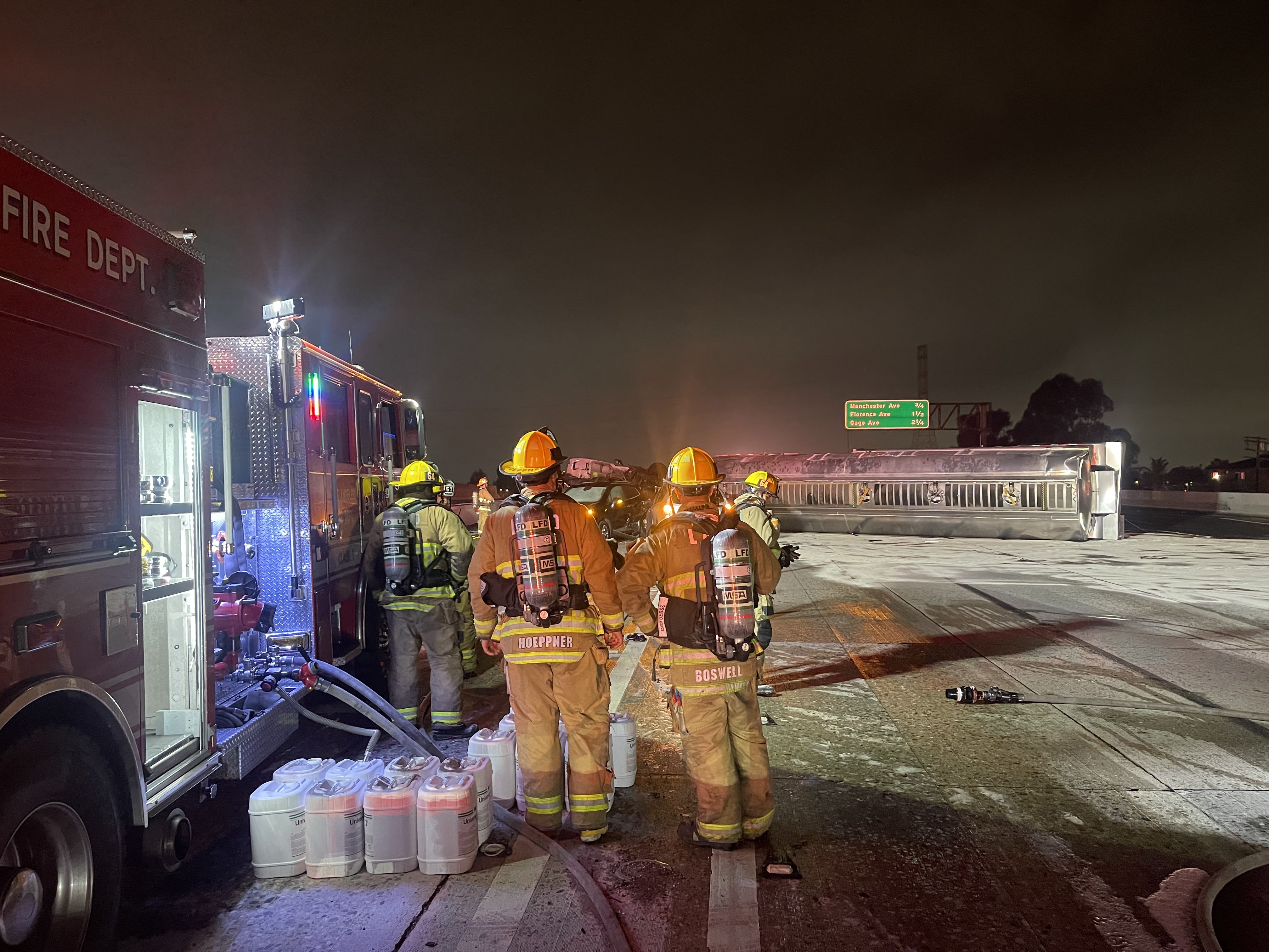 Foreground shows firefighters with equipment and overturned tank in background