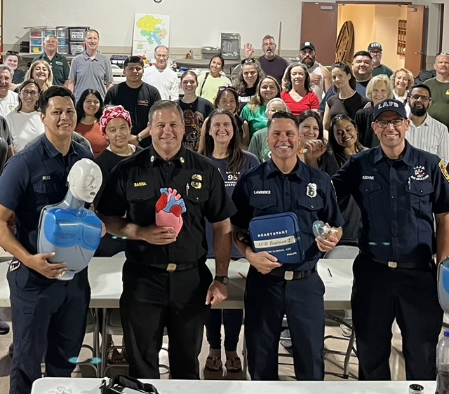 Four smiling LAFD personnel in the foreground hold training equipment—including a CPR manikin, an anatomical heart model, and an AED trainer case—while posing with a large, diverse group of smiling community members and volunteers in a classroom setting.