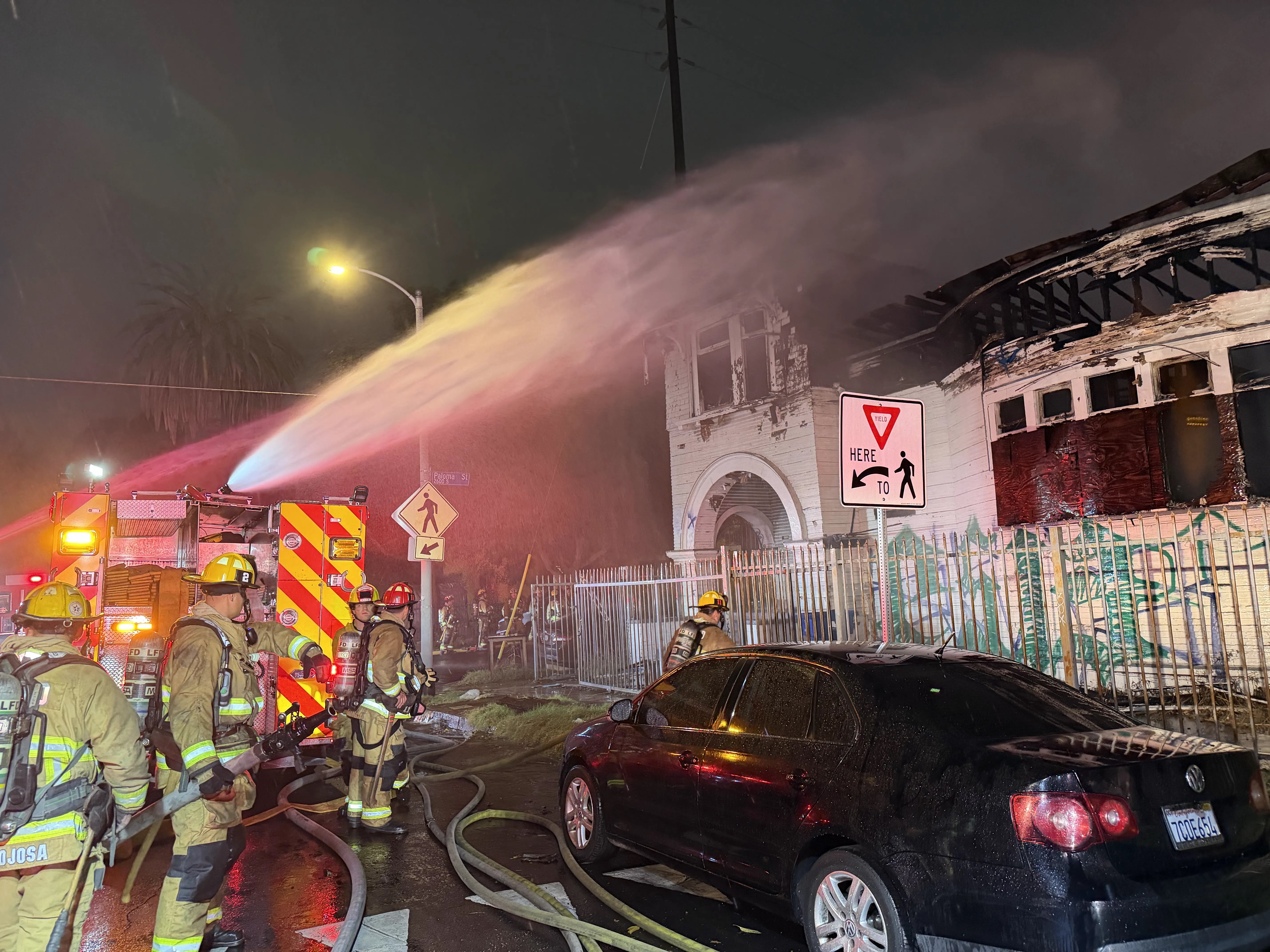 LAFD firefighters spray water onto a burning structure next to a street sign reading yield here to pedestrians